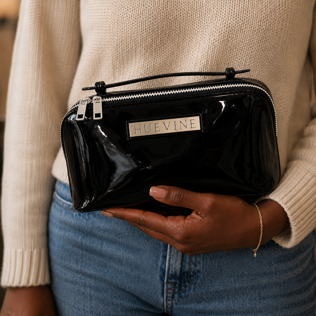 Person holding the HueVine Glow Bag black patent cosmetic case with silver zipper details and glossy finish.